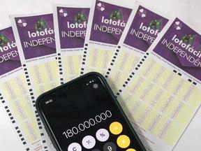 Cartelas da Lotofácil da Independência dispostas em fundo branco com celular em cima mostrando o número 180 mil