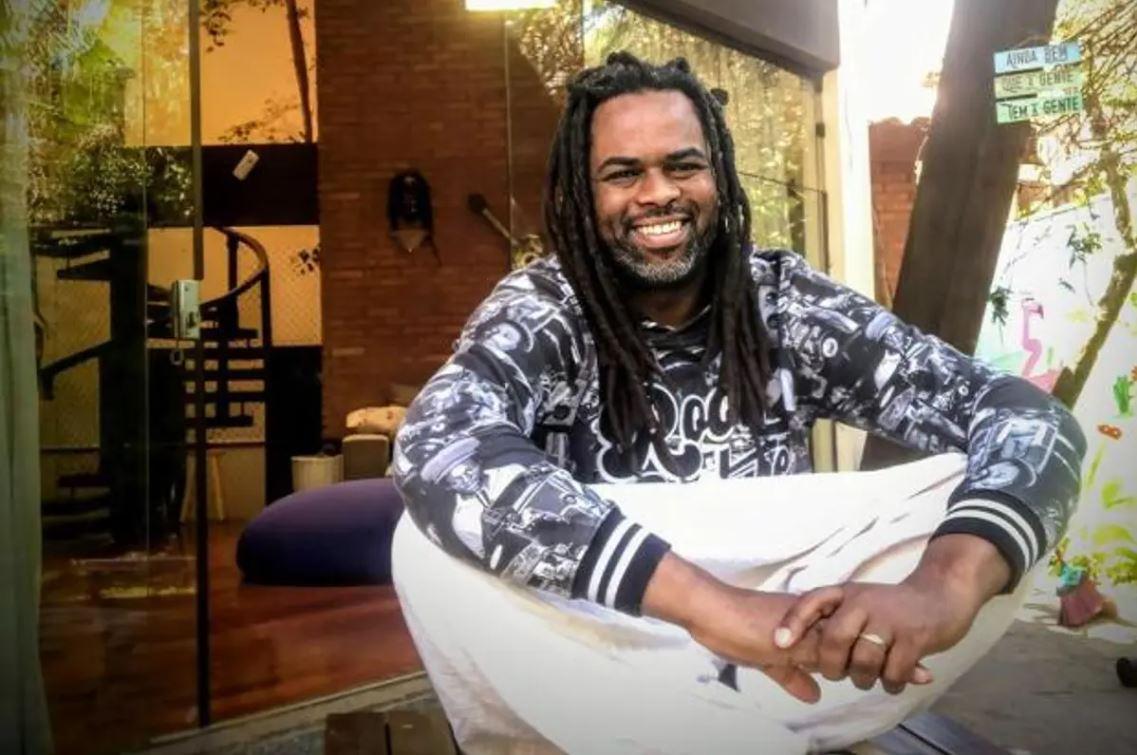 apresentador Manoel Soares, de blusa estampada e calça branca, sentado e sorrindo