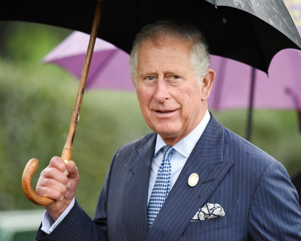 Charles é um homem branco e idoso, de cabelos grisalhos. Ele está de terno e segurando um guarda-chuva.