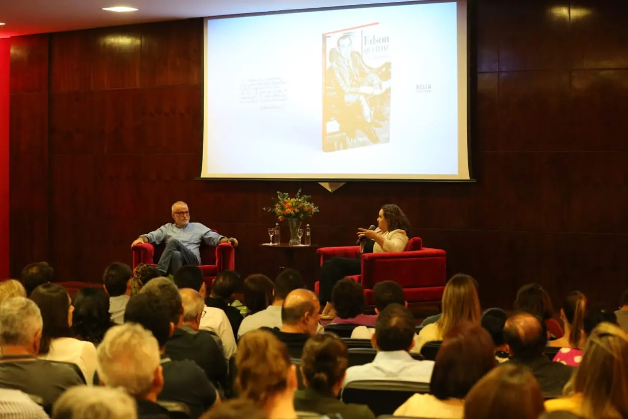 imagem mostra palco com telão projetando capa de livro atrás. lira neto está sentado à esquerda na foto com socorro acioli à direita, falando ao microfone
