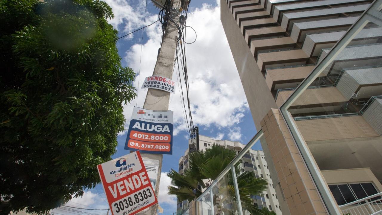 Ao fundo, um prédio alto. Na frente, placas de imobiliárias fixadas em postes dizendo : 'aluga