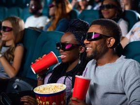Pessoas estão numa sala de cinema 3D. Em primeiro plano, um casal toma refrigerante e come pipoca. Eles são negros e usam óculos 3D.