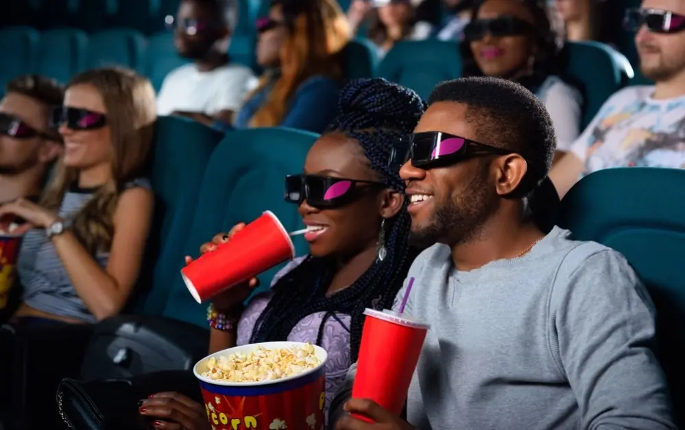 Pessoas estão numa sala de cinema 3D. Em primeiro plano, um casal toma refrigerante e come pipoca. Eles são negros e usam óculos 3D.
