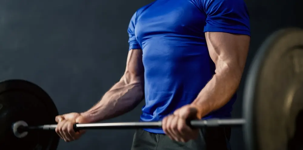 Homem segura barra com dois pesos. Ele veste uma blusa azul e um calção preto.