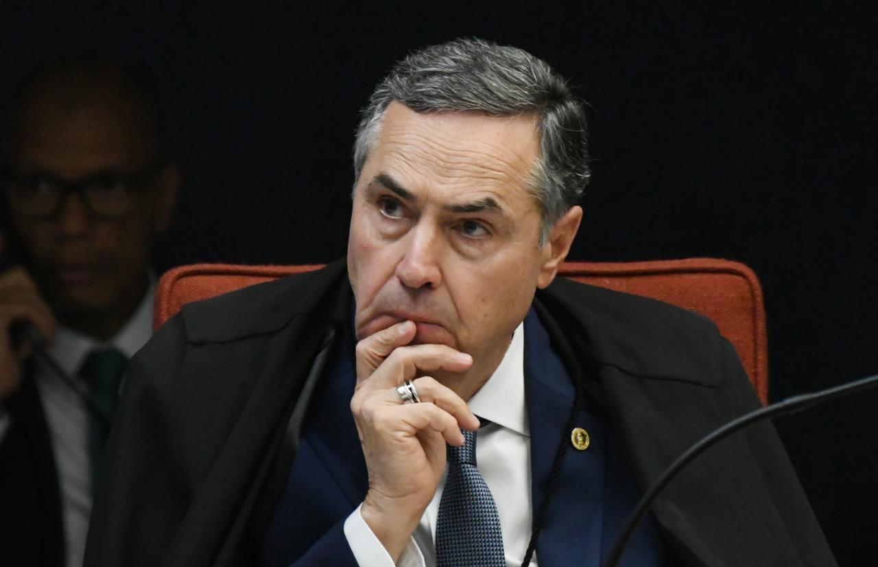 O ministro Roberto Barroso está com a mão no queixo, pensativo. Ele é um homem branco e tem o cabelo bem penteado para o lado.