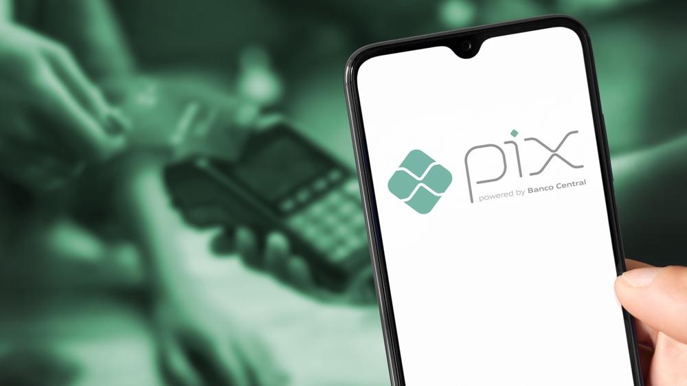 Imagem ilustrativa mostra um celular aberto no aplicativo do Pix. Em segundo plano, na cor verde, uma pessoa passa o cartão de crédito numa máquina.