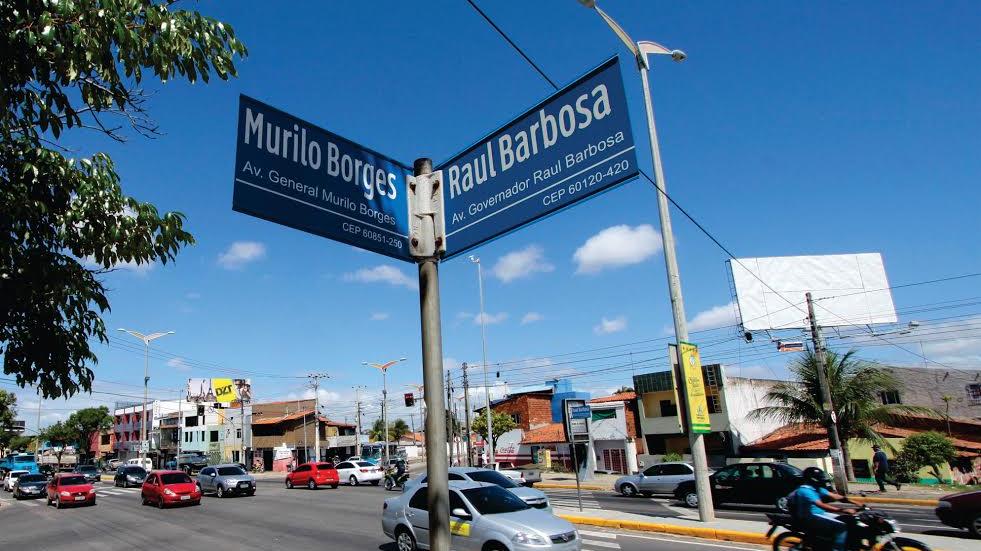Em primeiro plano, a imagem mostra as placas com os nomes das avenidas Murilo Borges e Raul Barbosa. Em segundo plano é possível observar carros e motocicletas trafegando pelo cruzamento