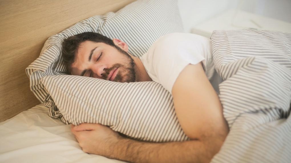 Homem branco e barbudo dormindo numa cama abraçado a um travesseiro.