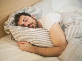 Homem branco e barbudo dormindo numa cama abraçado a um travesseiro.