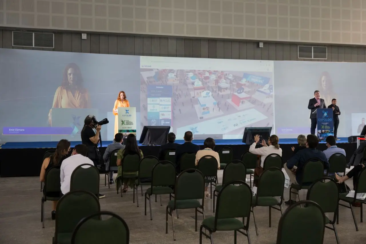 pessoas sentadas em cadeiras assistem a fala de duas pessoas em palco