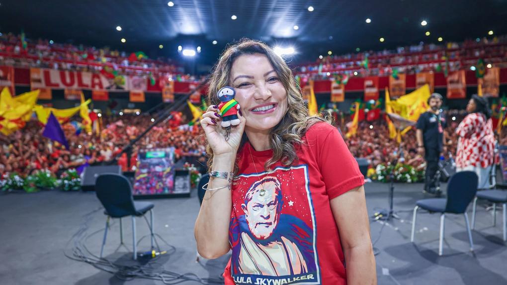 Janja está vestida com uma camisa vermelha com o rosto de Lula e segurando um bonequinho do presidente. Ela sorri diante de uma plateia cheia de apoiadores