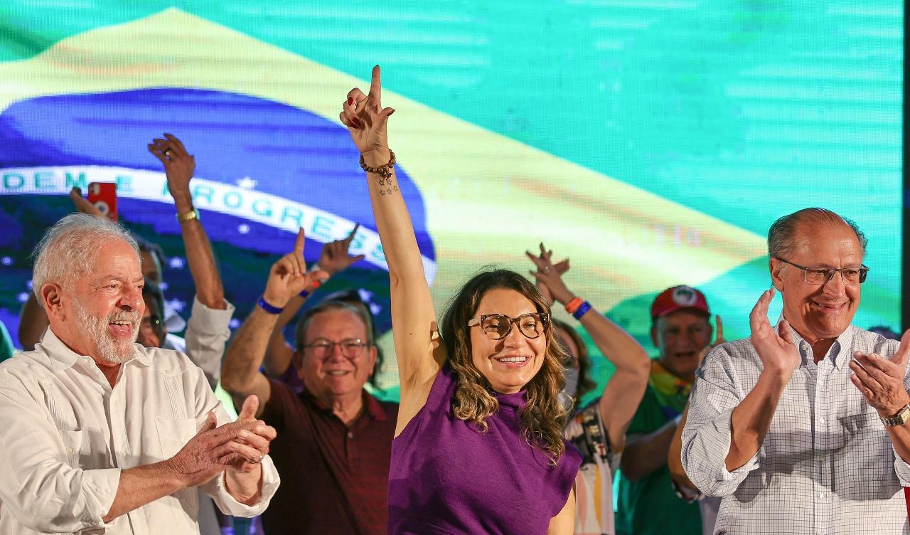 Lula está batendo palma à esquerda, Janja está vestida de roxo no centro fazendo o L e Alckmin está à direita batendo palmas