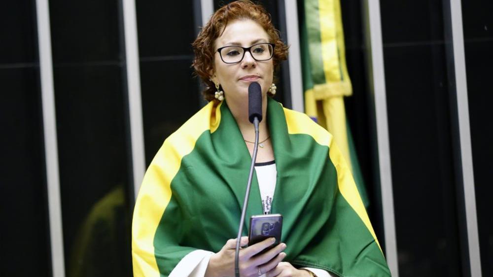 A deputada Carla Zambelli está com uma bandeira do Brasil sobre os ombros.