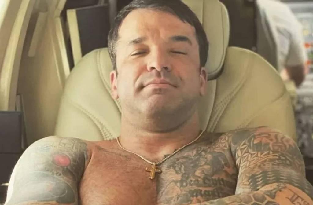 Thiago Brennand é um homem branco. Ele tem cabelo curto, muitas tatuagens pelo corpo e usa um escapulário dourado.