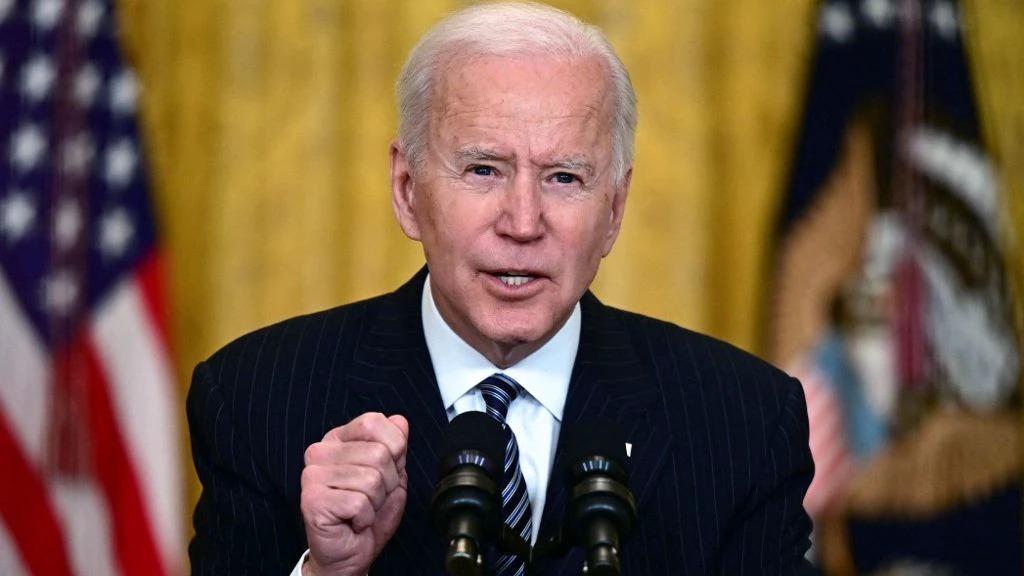 Joe Biden é um homem idoso, branco, de cabelos brancos e ralos. Ele está de terno preto, camisa branca e gravata azul listrada.