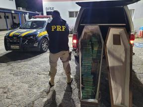 Na imagem é possível observar um agente da polícia rodoviária federal posando de costas ao lado de um caixão com 50 tabletes de crack, apreendido no interior de um carro funerária em 8 de novembro de 2022, em Vargem, Interior de São Paulo.