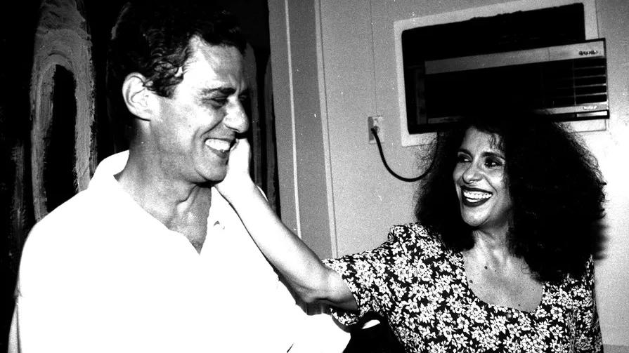 Foto em preto e branco mostra Gal Costa jovem com a mão no rosto de Chico Buarque. Os dois estão rindo