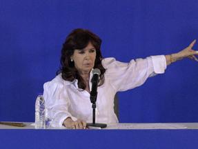 Cristina Kirchner é uma mulher idosa, de pele clara e cabelos pretos e lisos. Ela está vestindo uma blusa branca frouxa e de mangas compridas e com um braço esticado para o lado. Está falando ao microfone