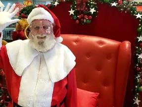 O ator Célio Figueiredo vestido como Papai Noel. Ele usa óculos transparentes, roupa de veludo vermelha com branco e uma barba branca