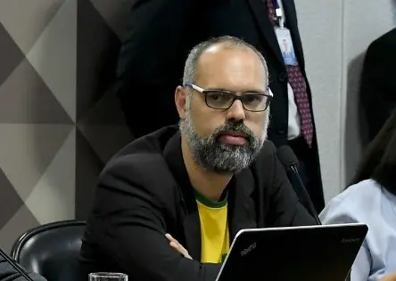 Alexandre dos Santos é um homem barbudo, calvo e usa óculos de armação preta.