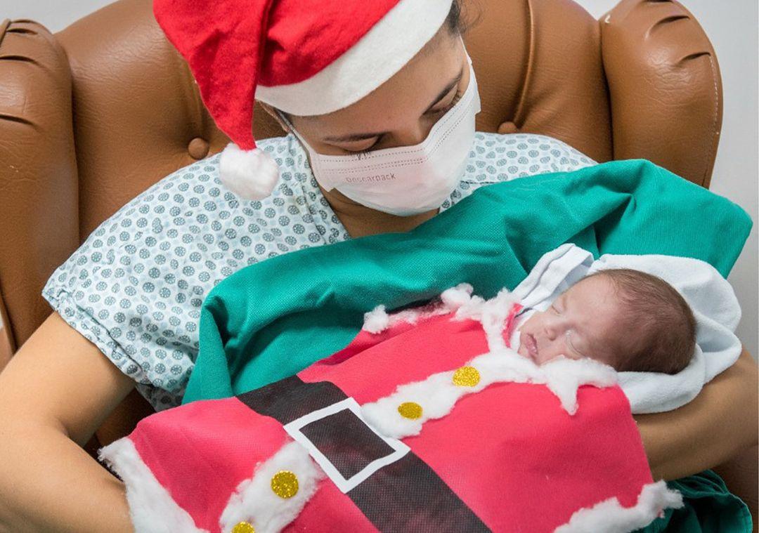 Paciente recém-nascido, internado na Unidade de Terapia Intensiva Neonatal (UTI Neo) do Hospital Geral Dr. Waldemar Alcântara (HGWA), posa ao lado da mãe usando roupas de temática natalina