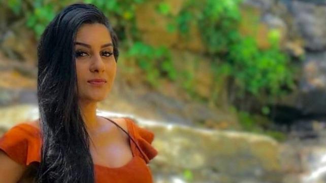 Samira tem longos cabelos escuros e está vestindo uma blusa vermelha com babados nos ombros