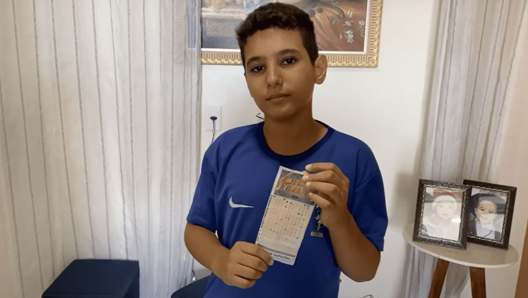 Menino de 10 anos, que acertou números sorteados na Mega da Virada, usando blusa azul e segurando bilhete do sorteio