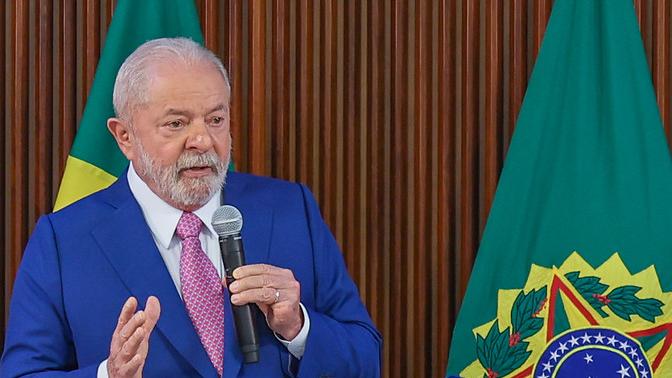 O presidente Lula está de terno azul e camisa branca e gravata rosa, segurando microfone