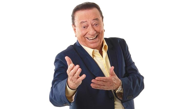 Raul Gil gesticula para a câmera e sorri. Fundo da foto é branco