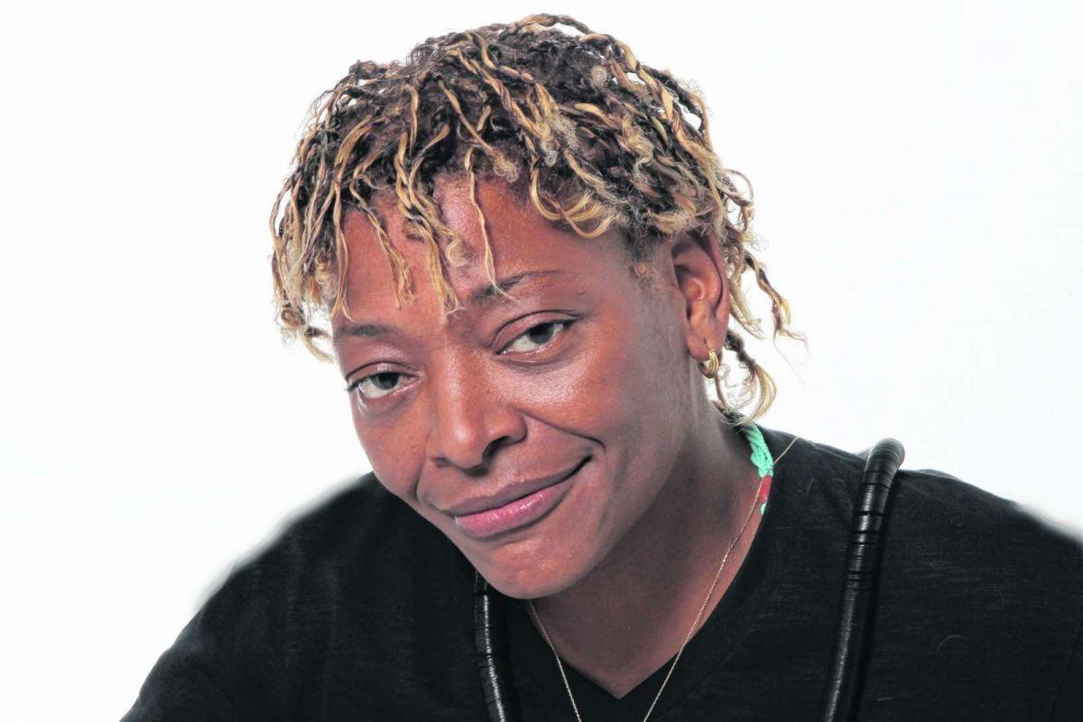 A cantora Mart'nália é uma mulher negra e de cabelos descoloridos. Ela está dando um sorriso tímido e o fundo da foto é branco