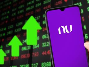 Imagem mostra um celular no aplicativo Nubank e símbolos de investimentos ao fundo