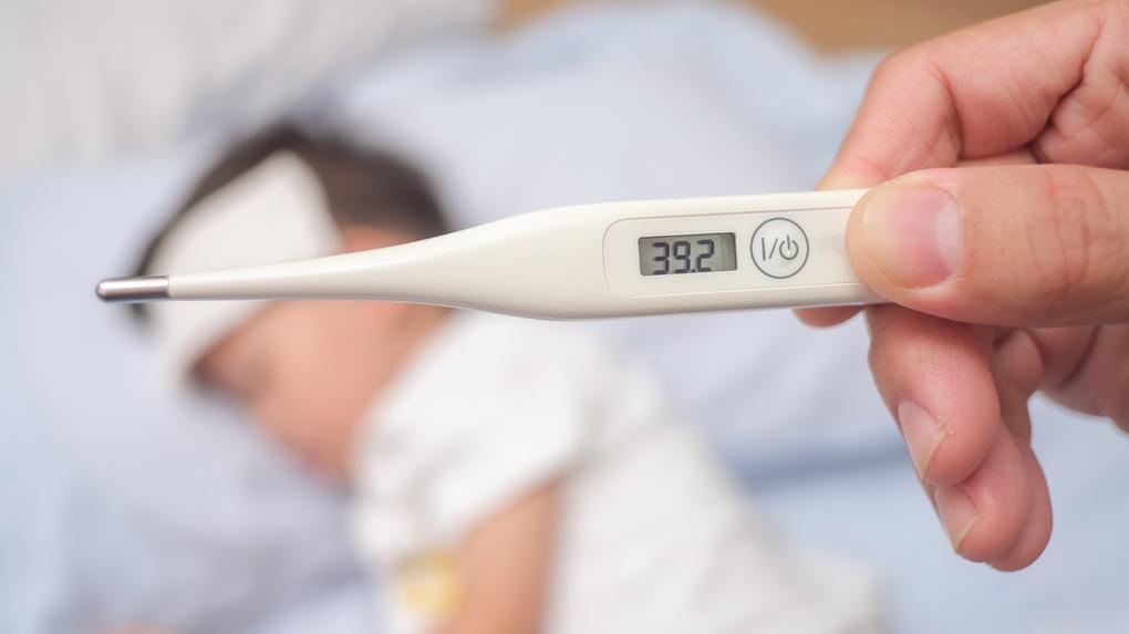Imagem mostram termômetro marcando temperatura de 39,2ºC. Ao fundo, uma criança deitada na cama, doente.