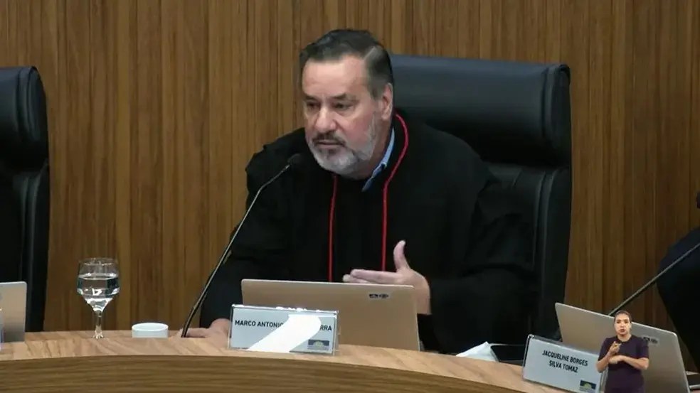 Marco é um homem branco, corpulento, de barba grisalha e cabelo escuro bem penteado.