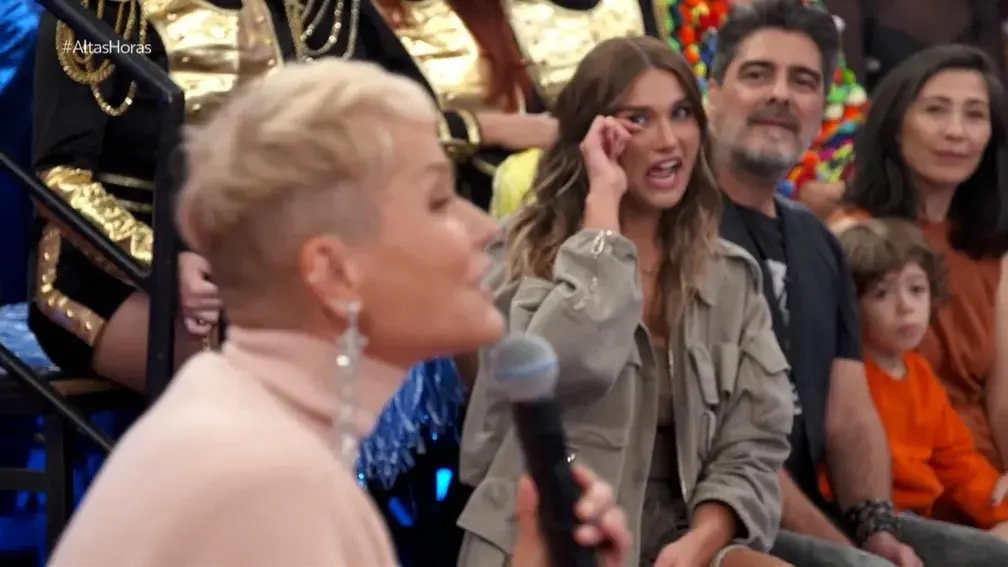 Xuxa fala ao microfone enquanto Sasha ouve e se emociona atrás, na plateia