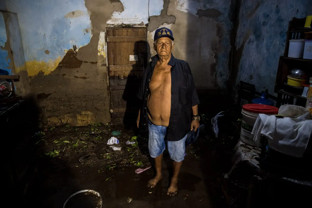 Francisco está de camisa de botão aberta e bermuda jeans. Ele usa um boné e sua expressão é de desesperança. Sua casa está totalmente destruída