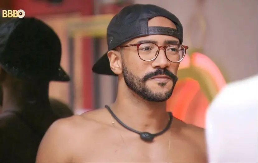 Ricardo é um homem negro de barba rala e bigode bem feito. Ele está sem camisa e usa óculos e boné para trás.