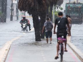 Avenida Historiador Raimundo Girão, em Fortaleza, alagada. Na foto, um motoqueiro, uma pessoa de bicicleta e pessoas a pé tentam passar pela água