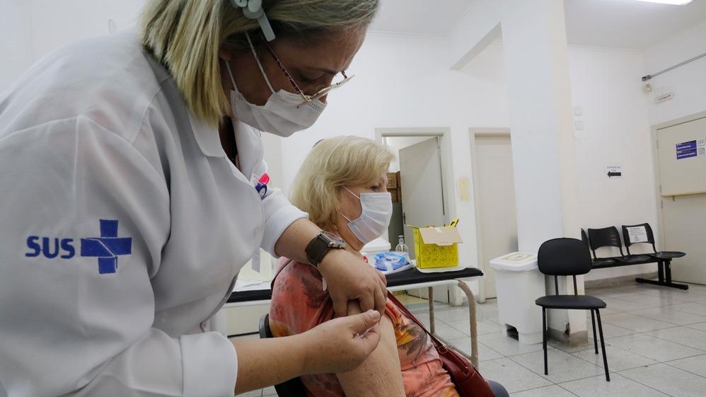 Médica aplica injeção em paciente idosa que está sentada num posto de saúde