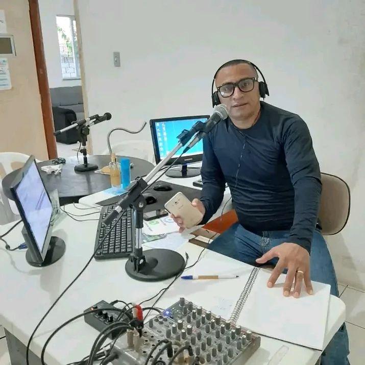 Gerson Ferreira é um homem negro, de pele clara, usa camisa azul com os braços cobertos e calça jeans. Ele está numa mesa de locução de rádio