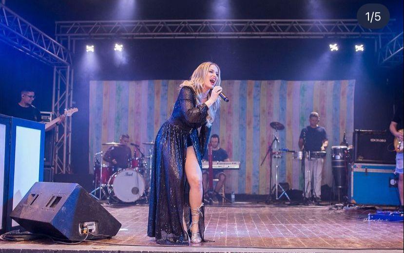 Nayara Vilela é uma mulher branca e loira. Ela está de vestido preto cantando segurando um microfone.