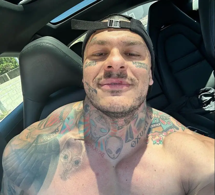 Toguro é um homem branco, corpulento, com o corpo repleto de tatuagens. Na foto, ele usa um boné preto para trás.