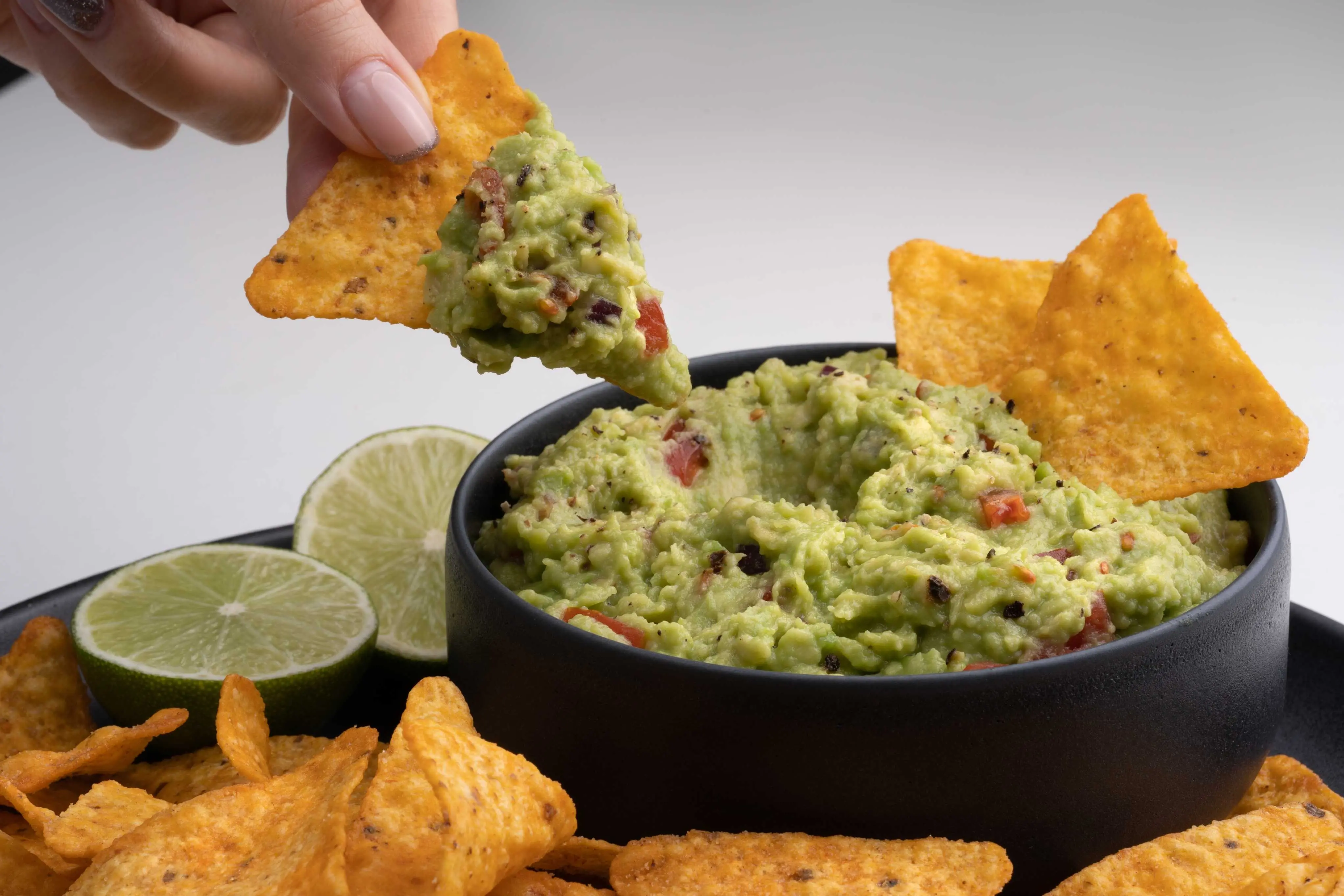 Imagem foca em mão mergulhando nacho em guacamole, ilustrando receita do prato