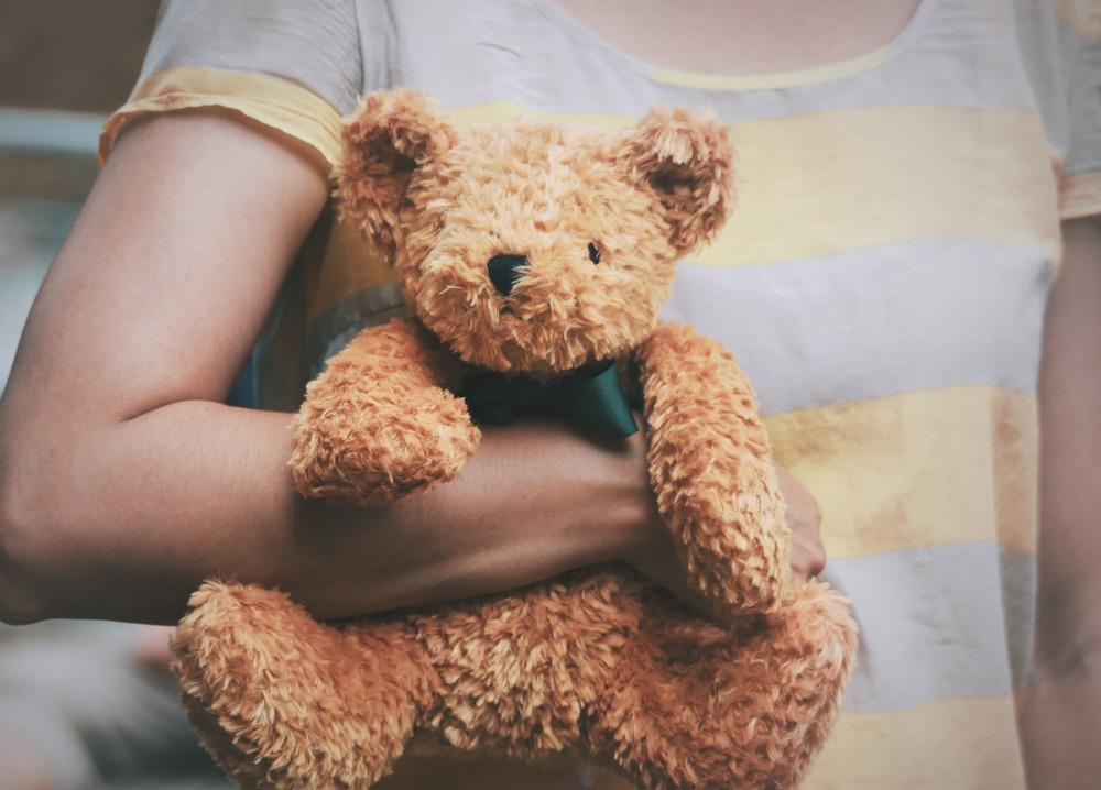 Criança sem identificação, de camisa listrada em amarelo e cinza, segura ursinho de pelúcia na cor marrom