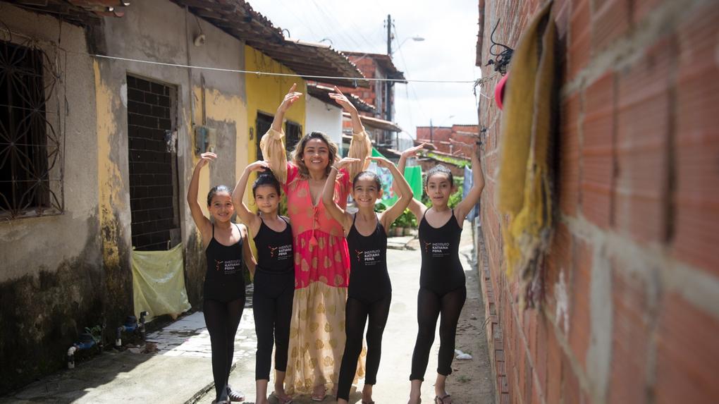 Mulher adulta e quatro meninas dançando balé em uma viela