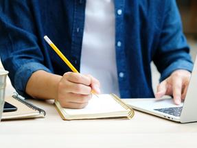 Homem digita em notebook enquanto escreve em um caderno com um lápis sobre uma escrivaninha