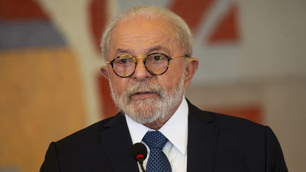 O presidente Lula discursa. Ele é um homem idoso, de cabelos e barbas brancos, e usa óculos de aros de tartaruga