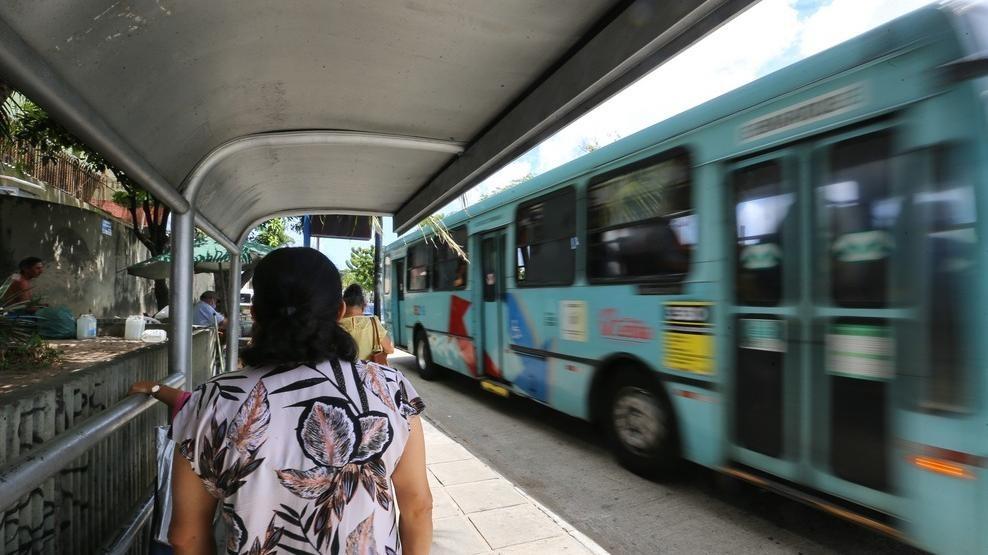 Mulher aguarda em ponto de ônibus enquanto um ônibus passa ao lado dela em alta velocidade