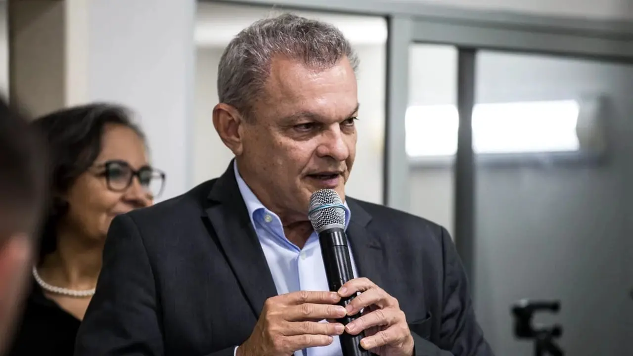 Sarto é um homem branco e de cabelos também brancos. Ele está de terno preto e camisa de botão azul, falando ao microfone