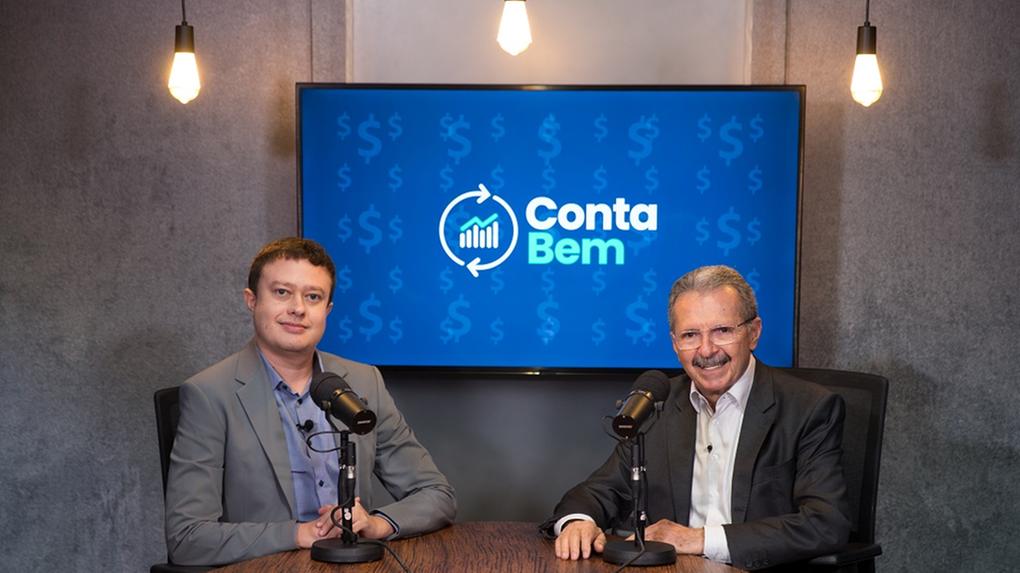 Dois homens sentados em uma mesa de apresentação de podcast. O da esquerda usa um terno cinza e o da direita um mais escuro