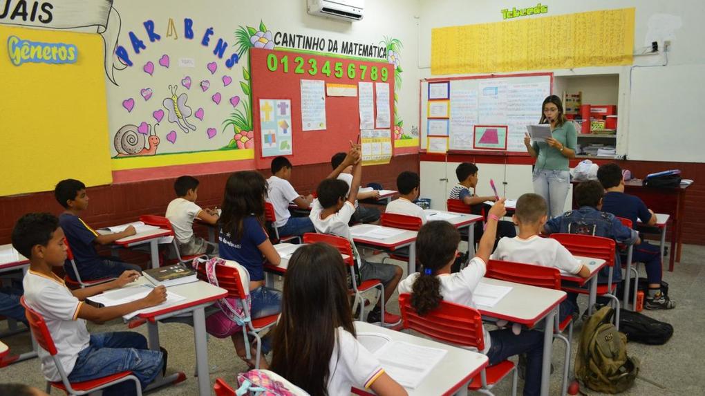 Sala de aula do ensino fundamental, com estudantes sentados e professora em pé, lendo